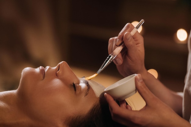 Um cosmetologista faz uma máscara para o rosto de uma mulher para rejuvenescer a pele Cosmetologia