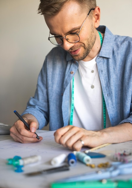 Um costureiro autodidata de óculos está trabalhando na mesa Um designer DIY desenha um esboço para um novo projeto de fantasia Um homem sorridente em uma camisa azul faz um esboço para costurar roupas como hobby