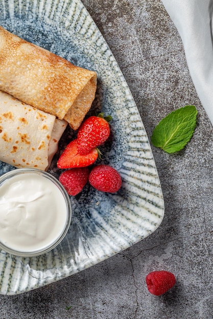 Um delicioso café da manhã. Panquecas doces fritas recheadas com morangos em um prato branco com creme de leite em uma mesa de pedra cinza com um guardanapo e talheres