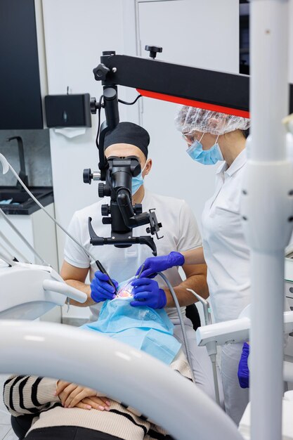 Um dentista de sucesso olha para os dentes do paciente com um microscópio odontológico e mantém instrumentos dentários perto da boca O assistente ajuda o médico Eles usam uniformes brancos com máscaras e luvas