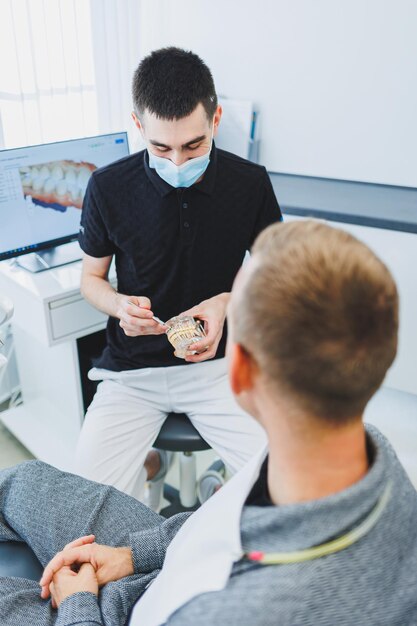 Um dentista em um consultório odontológico moderno conta a um paciente sobre cuidados dentários Um homem se senta em uma cadeira odontológica e ouve um dentista