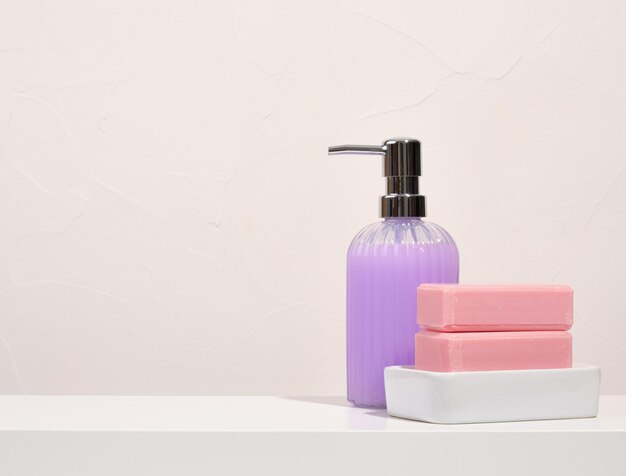 Um dispensador de gel de banho roxo e dois pedaços de sabonete rosa em uma saboneteira branca Copiar espaço para texto