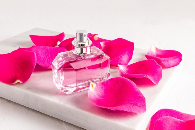 Um elegante frasco facetado de perfume feminino fica em um pódio de pedra branca de mármore com grandes pétalas de rosa chá rosa apresentação da fragrância