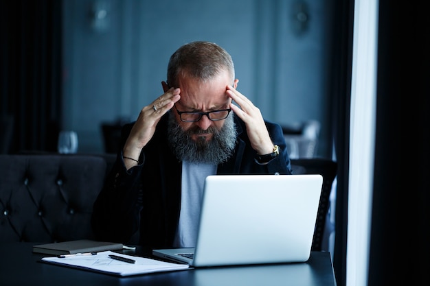 Um empresário adulto do sexo masculino está trabalhando em um novo projeto e olhando para os gráficos de crescimento das ações. senta-se à mesa perto da grande janela. olha para a tela do laptop e sorri