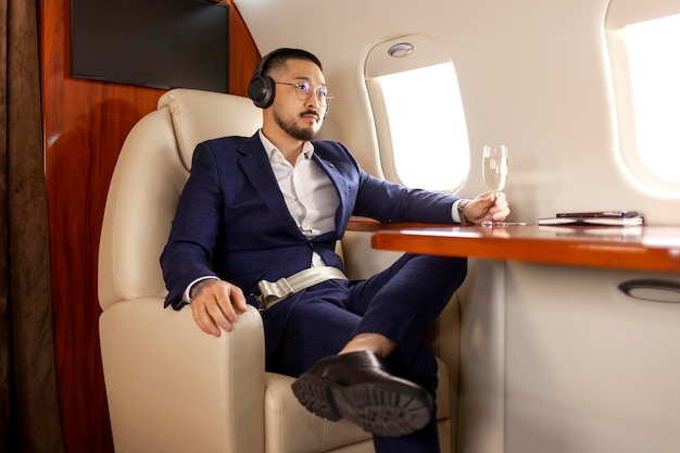 Um empresário asiático de sucesso voa num avião privado de luxo com um copo de champanhe e ouve música.
