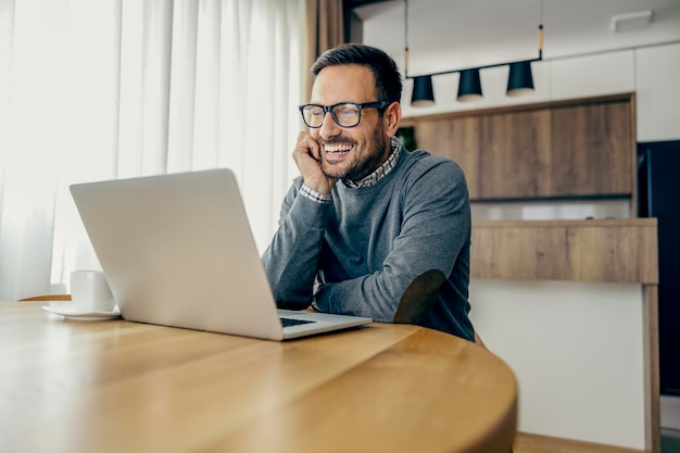 Um empresário casual sorridente está lendo e-mails no laptop enquanto trabalha remotamente em casa