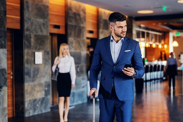 Um empresário de meia-idade vestido com roupas formais, caminhando no saguão de um hotel chique. ele está segurando um telefone inteligente e puxando uma mala.