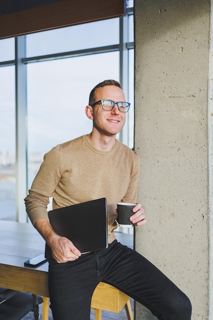 Um empresário de óculos e roupas elegantes bebe café em um espaço de trabalho moderno Um freelancer masculino com um laptop trabalha remotamente