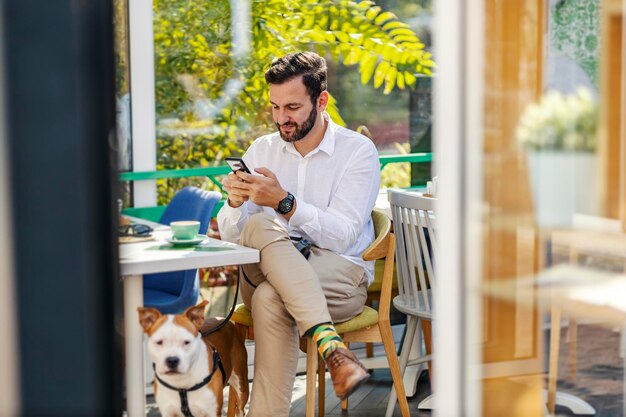 Um empresário está sentado em um café que aceita animais de estimação com seu cachorro e digitando mensagens nas redes sociais
