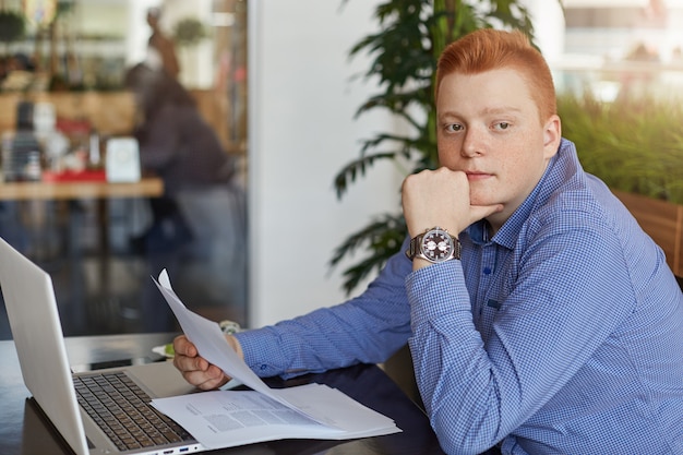 Um empresário ruivo jovem pensativo vestindo camisa formal, sentado à mesa com o laptop e documentos