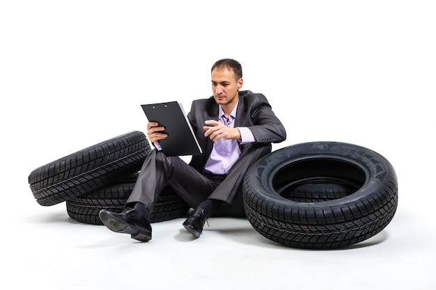 Um empresário sério sentado sobre uma pilha de pneus de carro. Oficina. Serviço automotivo. Revenda de peças sobressalentes. solado em fundo branco