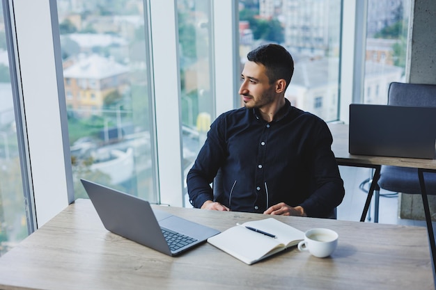 Um empresário sorridente em uma camisa preta está sentado em uma mesa usando um laptop Uma pessoa trabalha no escritório em um computador analisa dados Marketing na Internet educação de adultos elearning