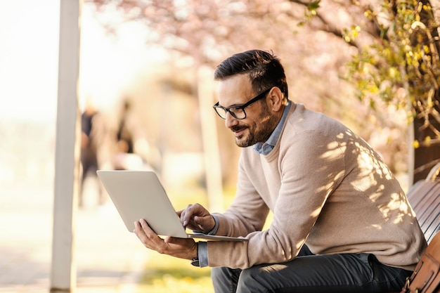 Um empresário urbano está sentado no banco do parque e digitando em seu laptop