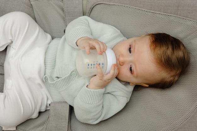 Um encantador bebê de olhos azuis de 5 meses deita-se na cama e bebe leite de uma mamadeira com uma mamadeira