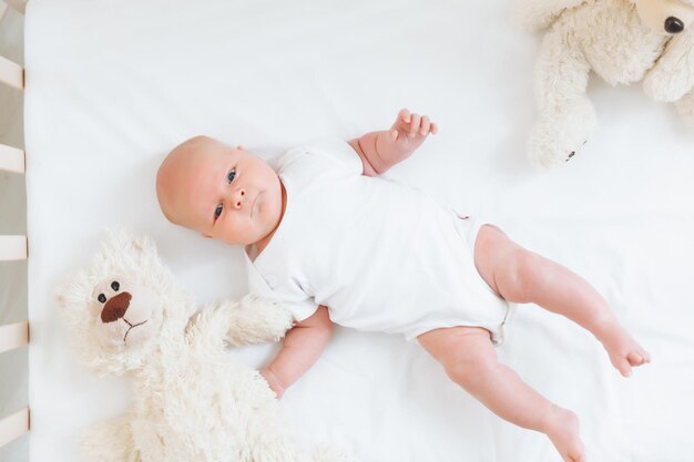 Um encantador e sorridente bebê de 2 meses de idade em uma roupa branca está em um berço ao lado de um ursinho de pelúcia vista de cima de um recém-nascido