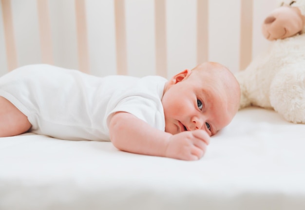 Um encantador e sorridente bebê de 2 meses de idade em uma roupa branca está em um berço ao lado de um ursinho de pelúcia Vista de cima de um recém-nascido