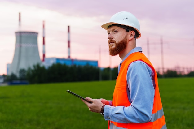 Um engenheiro com um tablet no fundo de uma usina de energia Inspeção de sistemas de engenharia Um homem de capacete e colete laranja