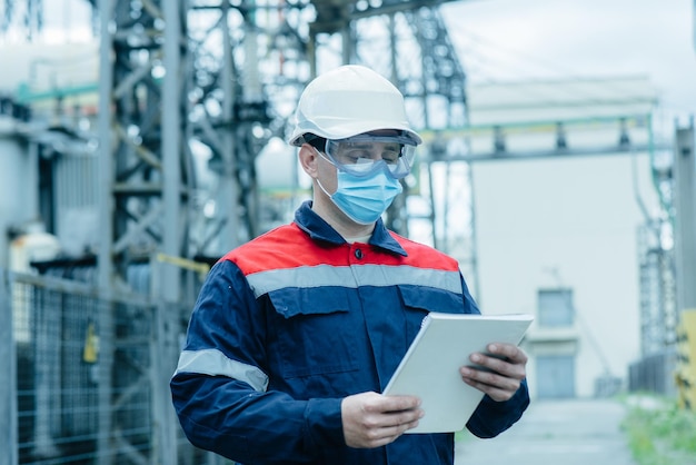 Foto um engenheiro de energia mascarado durante uma pandemia inspeciona o equipamento moderno de uma subestação elétrica antes do comissionamento energia e indústria reparo programado de equipamentos elétricos