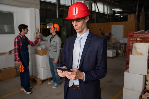 Um engenheiro usa um tablet no canteiro de obras enquanto usa um capacete e os trabalhadores atrás dele estão trabalhando