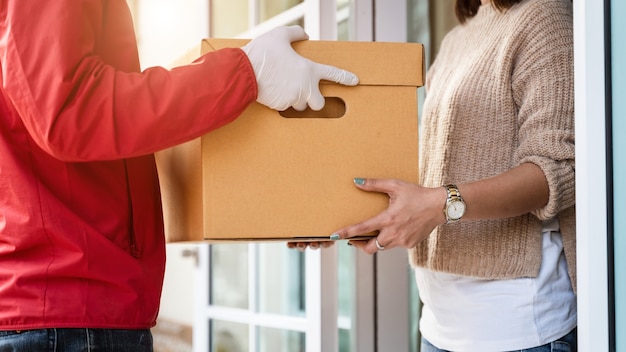 Foto um entregador asiático em uniforme vermelho entregando um pacote a uma cliente em frente à casa. um carteiro e um serviço de entrega expressa entregam o pacote durante a pandemia de covid19.