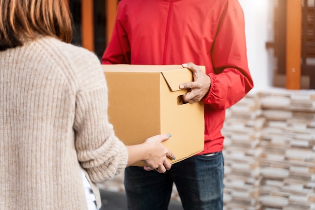 Um entregador asiático em uniforme vermelho entregando um pacote a uma cliente em frente à casa. Um carteiro e um serviço de entrega expressa entregam o pacote durante a pandemia de covid19.