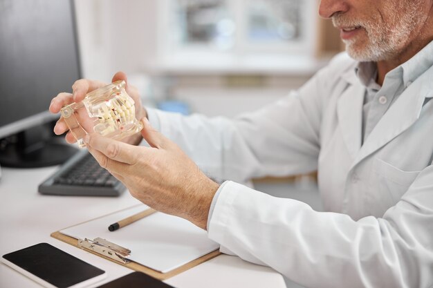 um especialista dentário idoso olhando para uma prótese dentária enquanto está sentado em sua mesa