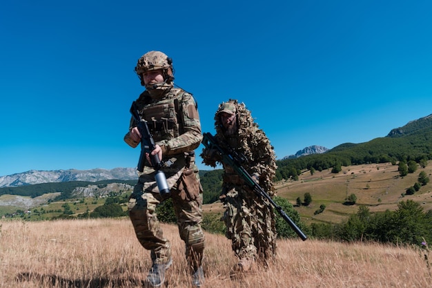 Um esquadrão de soldados da equipe de atiradores está disfarçado. Assistente de atirador e líder de equipe caminhando e mirando na natureza com grama amarela e céu azul. Uniforme de camuflagem tática.