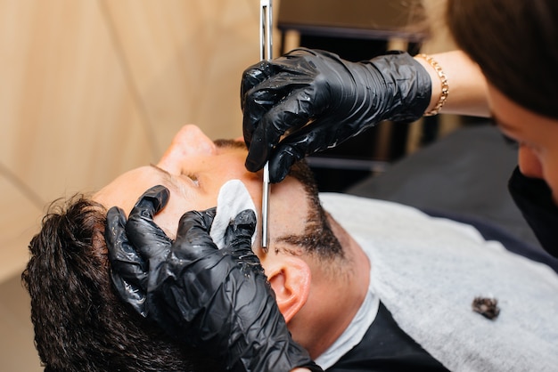 Um estilista profissional em uma barbearia moderna e estilosa faz a barba e corta o cabelo de um jovem. Salão de beleza, salão de cabeleireiro.