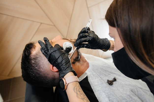 Um estilista profissional em uma barbearia moderna e estilosa faz a barba e corta o cabelo de um jovem. Salão de beleza, salão de cabeleireiro.