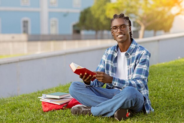 Um estudante afro-americano feliz lê um livro ao ar livre.