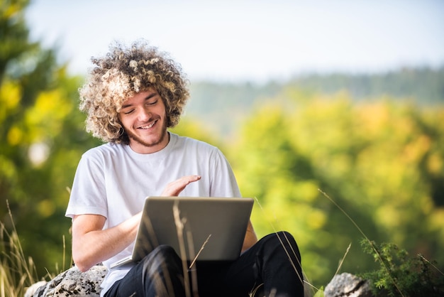 Um estudante com um penteado afro sentado na natureza e usa um laptop durante uma pandemia de vírus corona