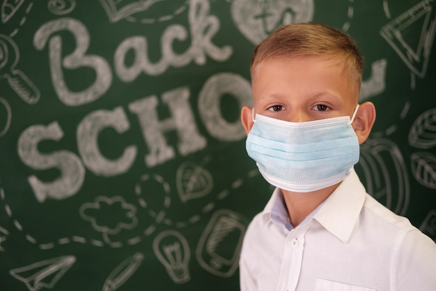 Foto um estudante com uma máscara médica no fundo de uma lousa com o texto de volta às aulas