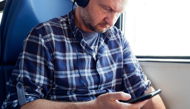 Um europeu de 40 anos com uma camisa xadrez azul está sentado em um trem ouvindo música em seus fones de ouvido