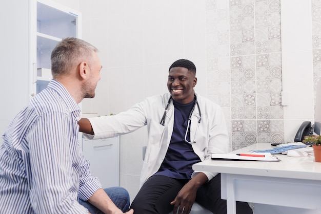 Um feliz e sorridente médico afro-americano fala com um paciente enquanto está sentado em uma mesa