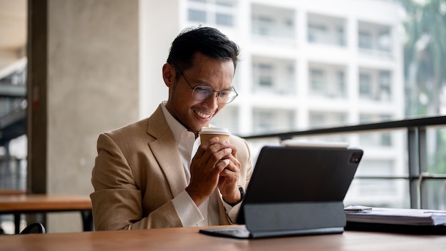 Foto um feliz empresário asiático sorri enquanto segura uma xícara de café descartável enquanto olha para um tablet