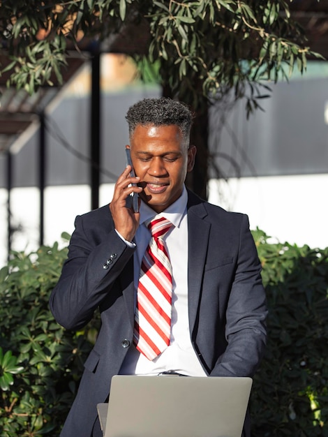 Um feliz empresário hispânico sorridente falando em um smartphone e trabalhando remotamente em seu laptop