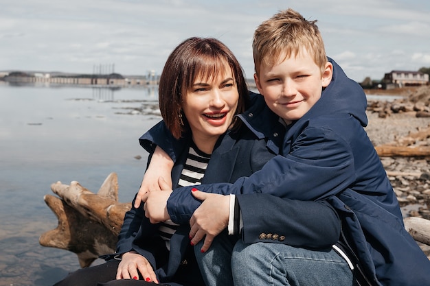 Um filho abraça sua mãe na margem do rio. Uma mãe bonita e moderna com um filho feliz sentado sobre troncos.