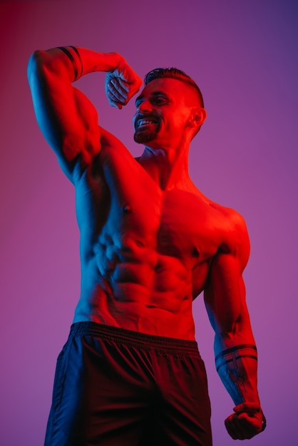 Um fisiculturista sorridente com barba posa com o bíceps flexionado sob a luz azul e vermelha