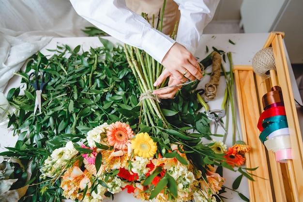 Um florista amarra um buquê de flores frescas com barbante em sua mesa cheia de folhas cortadas