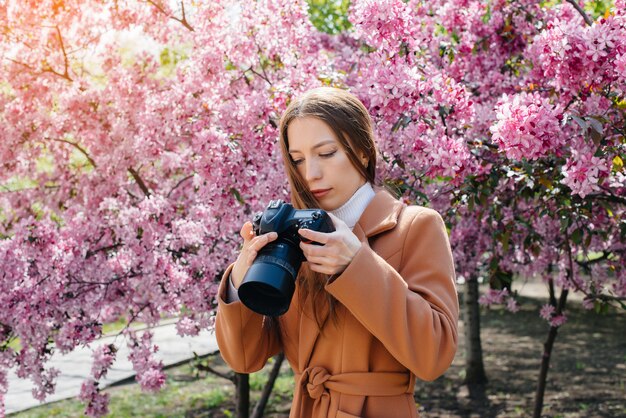 Um fotógrafo jovem bonita caminha e tira fotos contra uma macieira florescendo. hobbies, recreação.