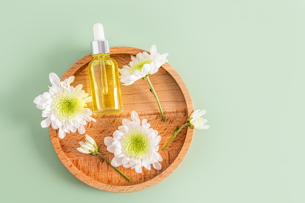 Um frasco cosmético com uma pipeta com um produto orgânico para cuidados com a pele facial está sobre uma placa de madeira com fundo verde de vista superior de flores