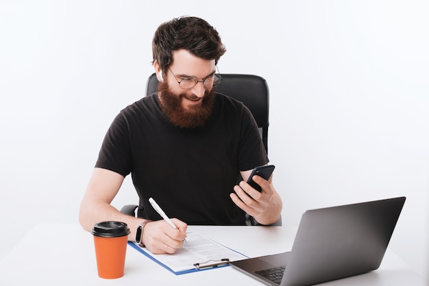 Um freelancer barbudo trabalhando, fazendo seu trabalho de papel e usando telefone celular sobre o espaço isolado