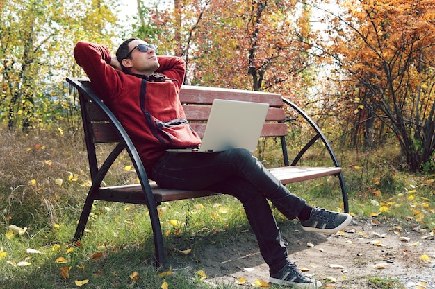 Um freelancer de funcionário remoto no fundo do outono Park Jovem empresário está sentado no parque e usando engenheiro de software de laptop usando laptop ao ar livre Cara relaxado descansando no parque depois do trabalho