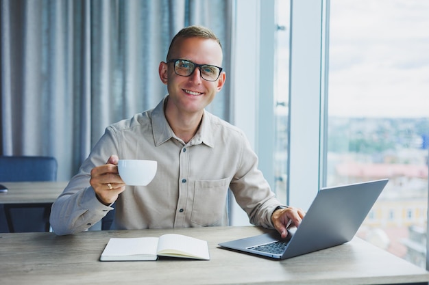 Um freelancer de óculos trabalha em um laptop e bebe café um gerente se senta em uma mesa no escritório trabalha em um laptop Funcionário freelancer no local de trabalho no trabalho remoto