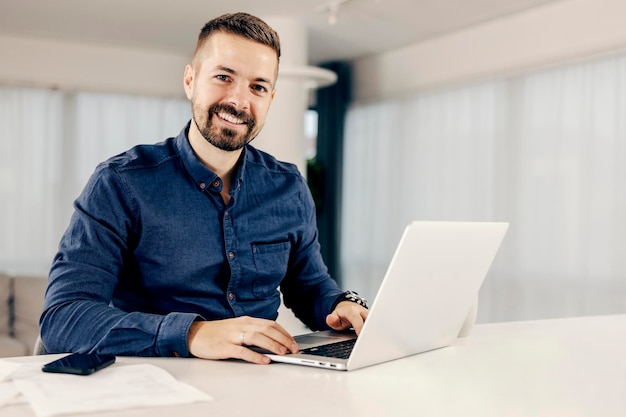 Um freelancer feliz trabalhando em sua casa aconchegante no laptop e sorrindo para a câmera