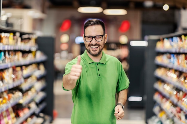 Foto um funcionário feliz está parado no supermercado entre os corredores e dando o polegar para cima