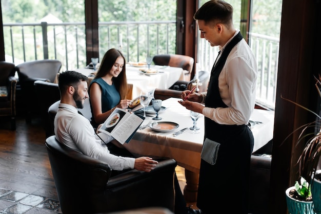 Um garçom elegante serve um jovem casal de um homem e uma mulher que veio a um restaurante gourmet Atendimento ao cliente no restaurante e estabelecimentos de catering