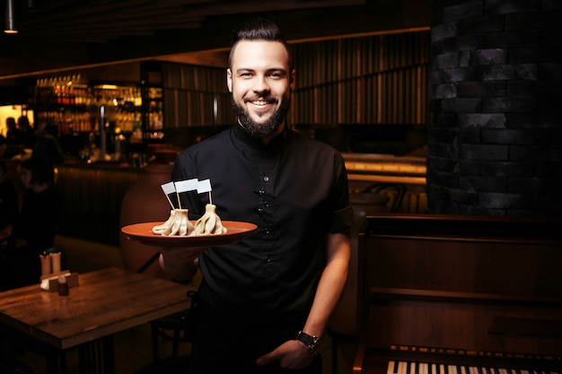 Foto um garçom exigente em roupas pretas, com barba e uma porção de khinkali