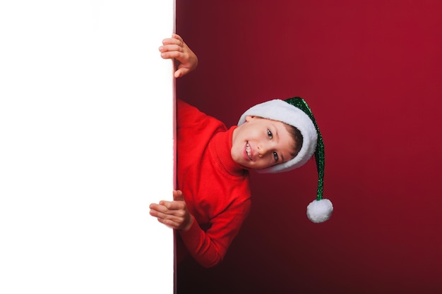 Foto um garotinho com um chapéu de papai noel posando atrás de um painel branco isolado em um fundo vermelho uma criança bonita está segurando um outdoor de natal vazio uma criança espia o quadro para os feriados