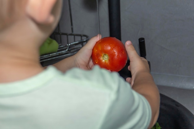 Um garotinho com um corte de cabelo curto ajuda a cozinhar na cozinha Lava legumes frescos e ervas em uma pia preta em uma cozinha cinza